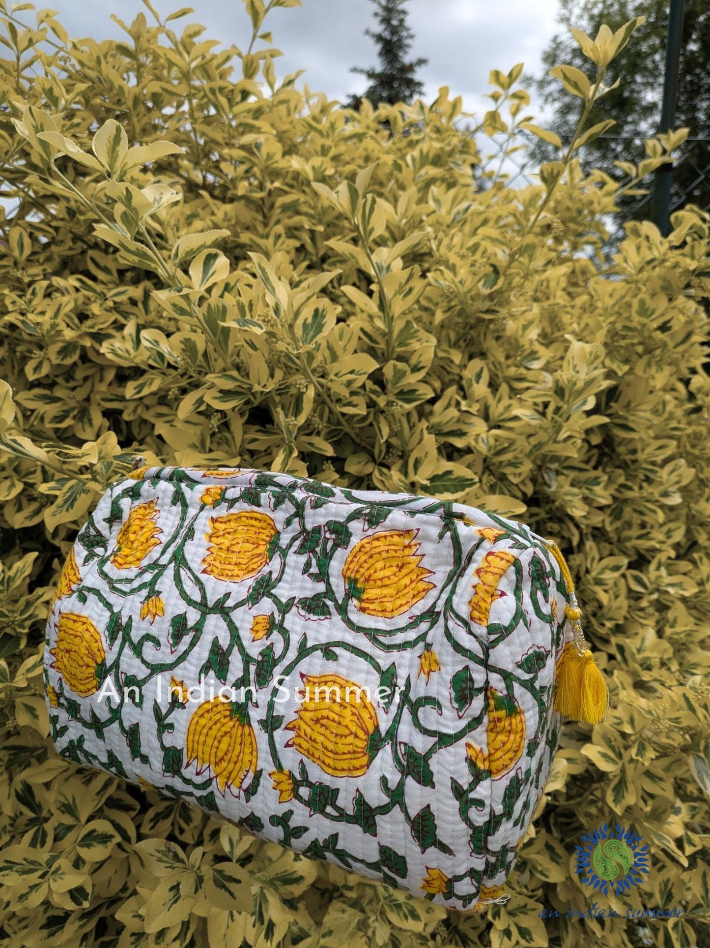 A yellow washbag with a floral hand block print and beaded tassels, displayed against a natural background with green leaves.
